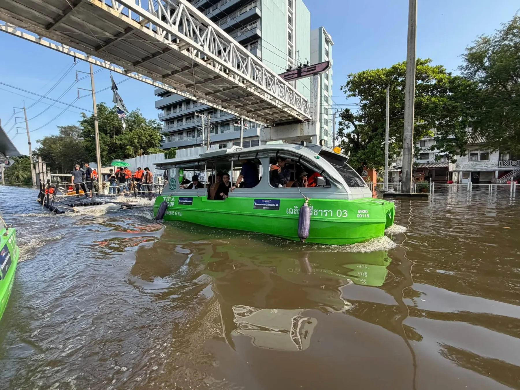 เรือไฟฟ้าสีเขียวบนถนนที่น้ำท่วม ใต้สะพานลอยคนเดินข้าม