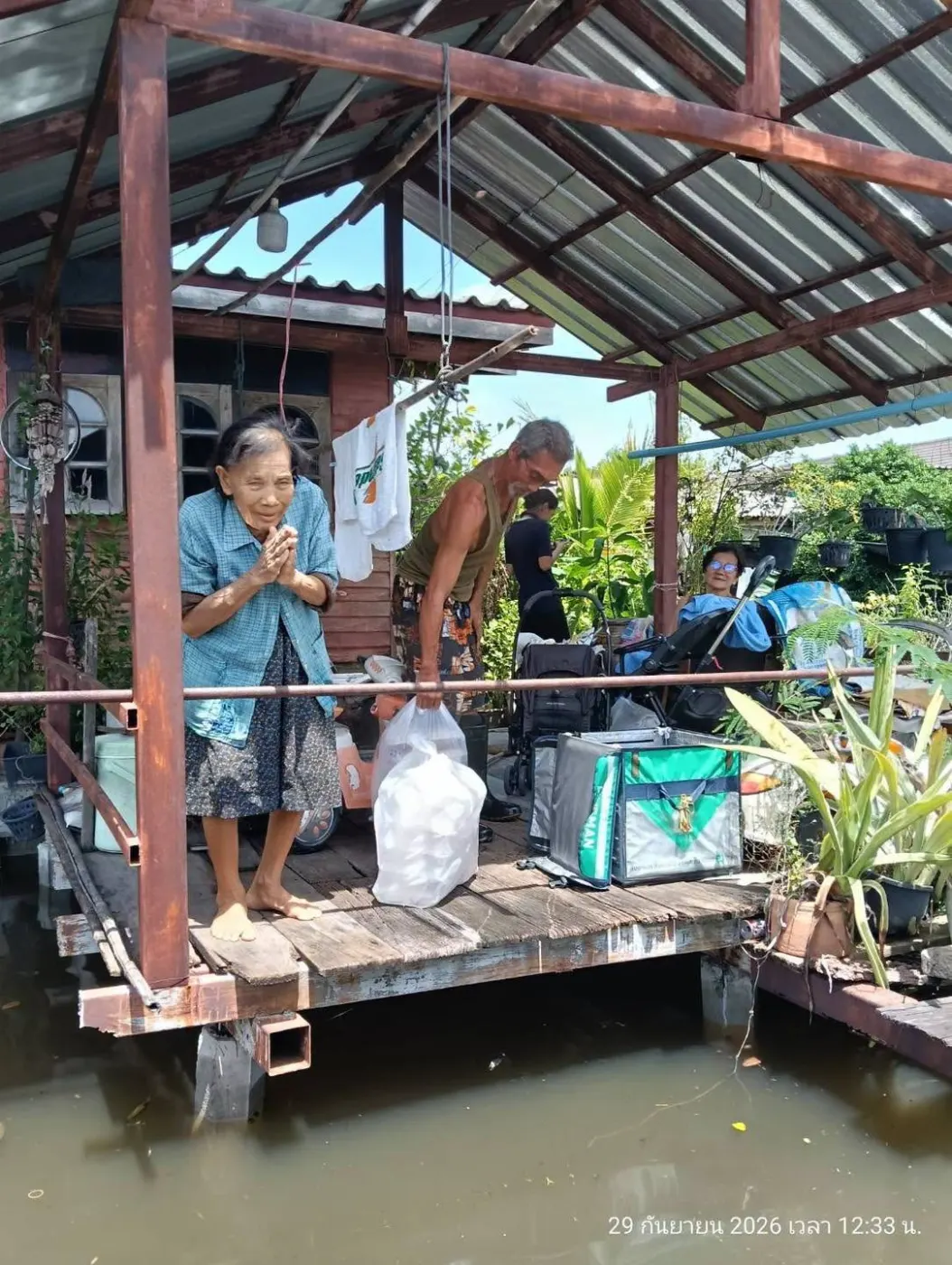 ผู้สูงอายุบนระเบียงบ้านที่ถูกน้ำท่วม พร้อมถุงสิ่งของบรรเทาทุกข์